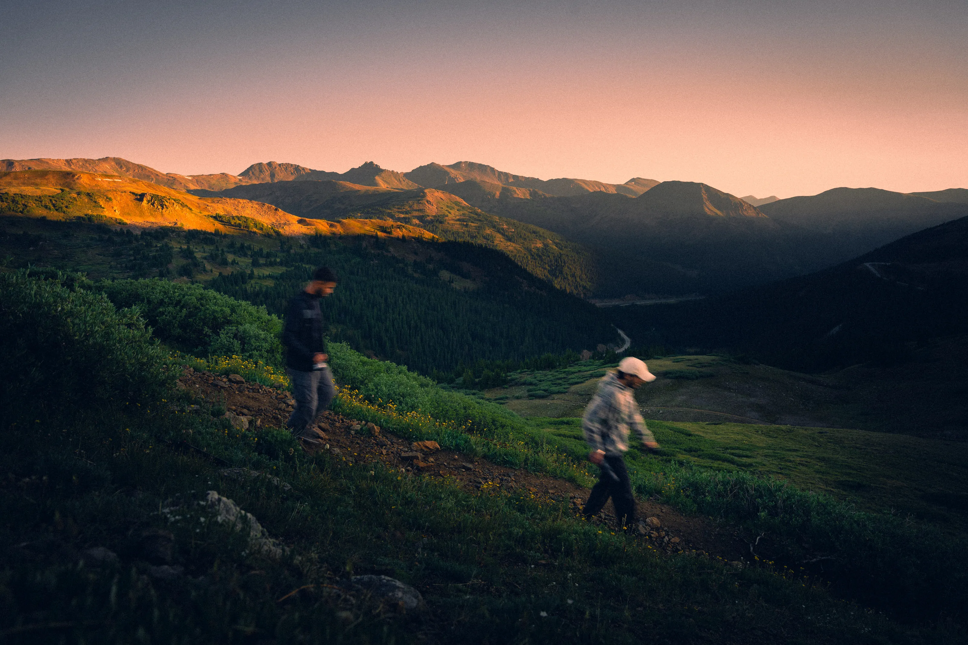 Two hikers cross a green slope as the last light turns the ridgeline orange.