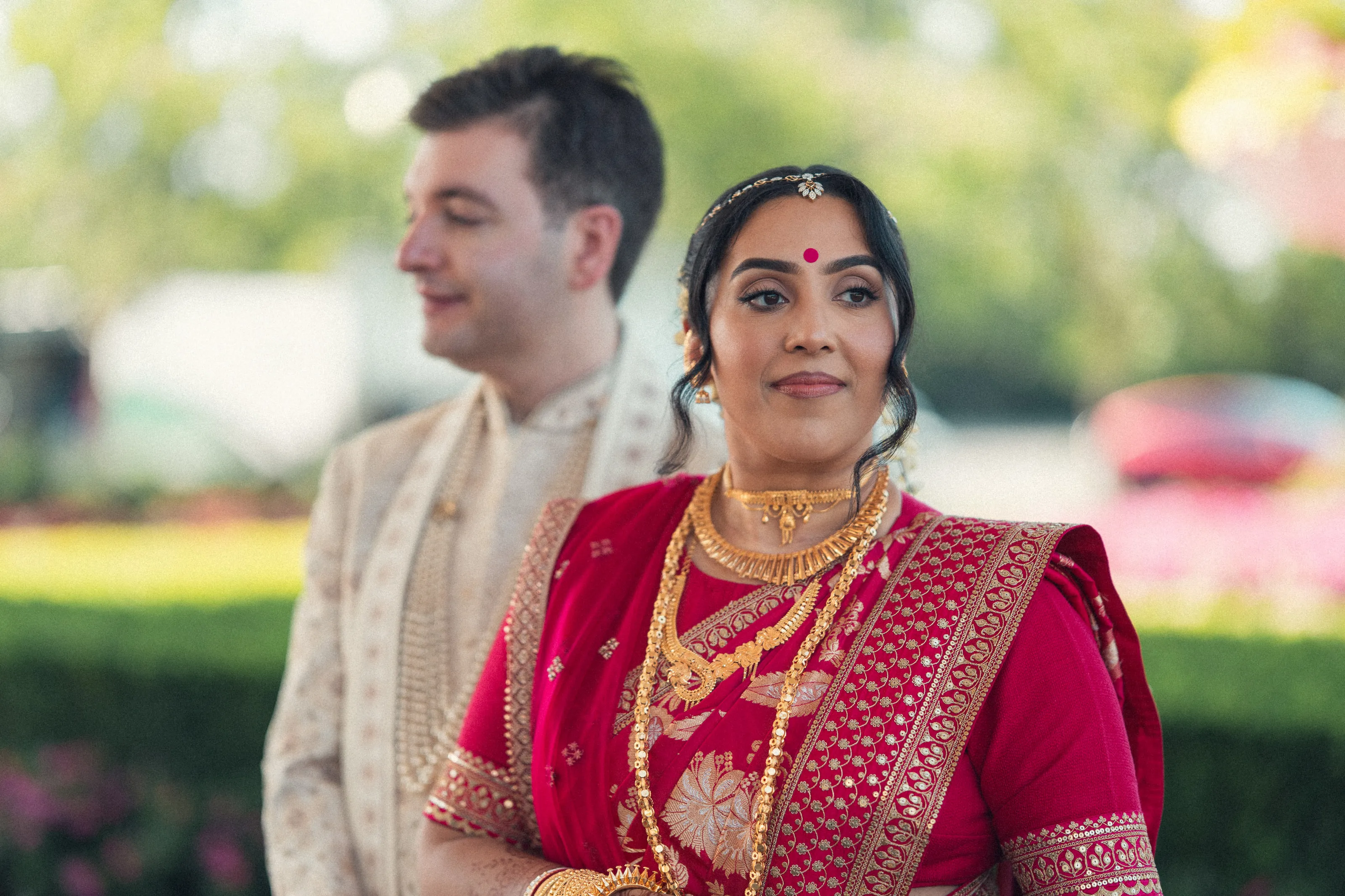 A bride in a red sari and gold jewellery stands beside the groom in a cream sherwani, in a garden.