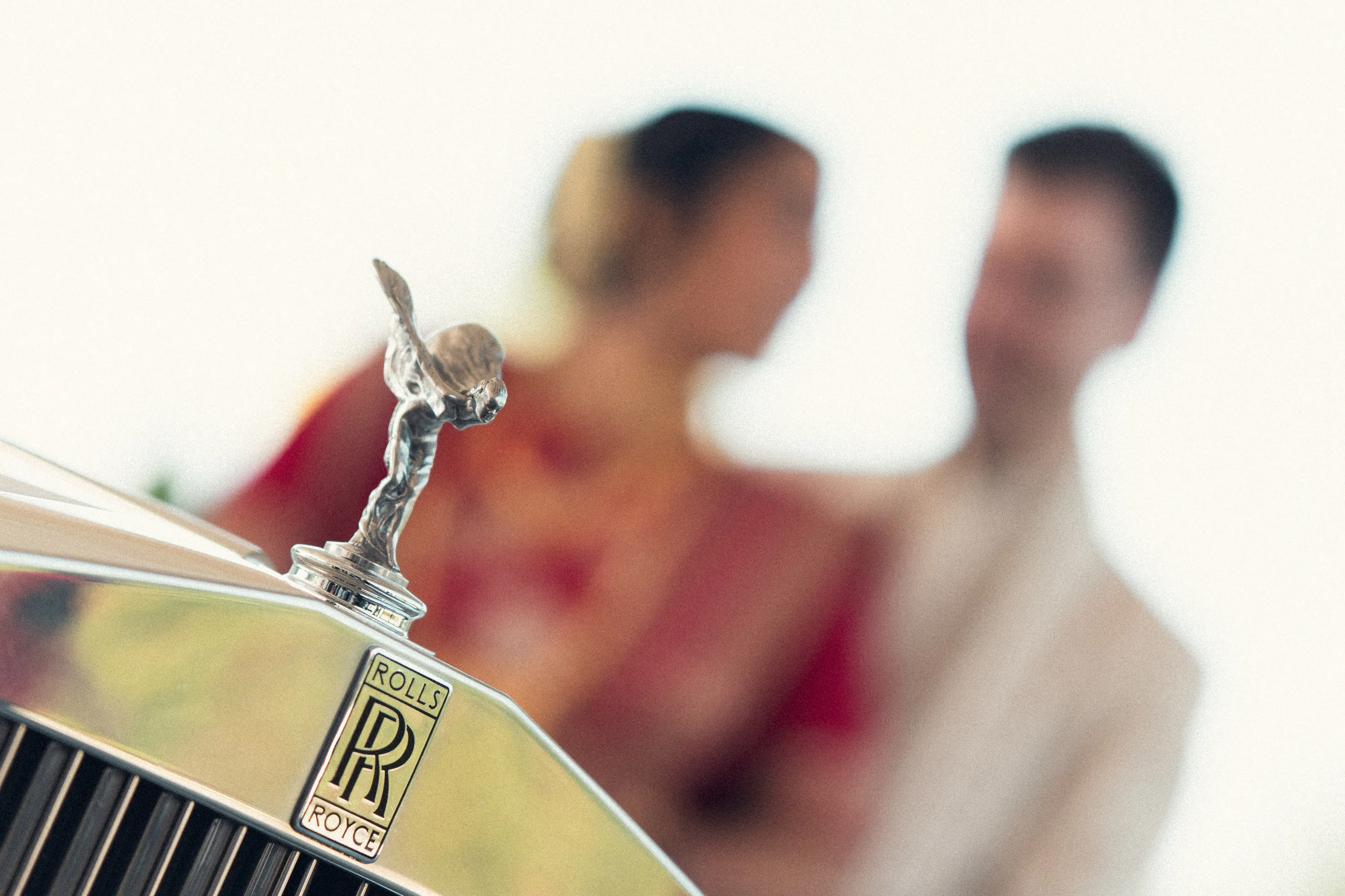 A silver Rolls-Royce mascot in sharp focus, with the bride and groom blurred behind it.