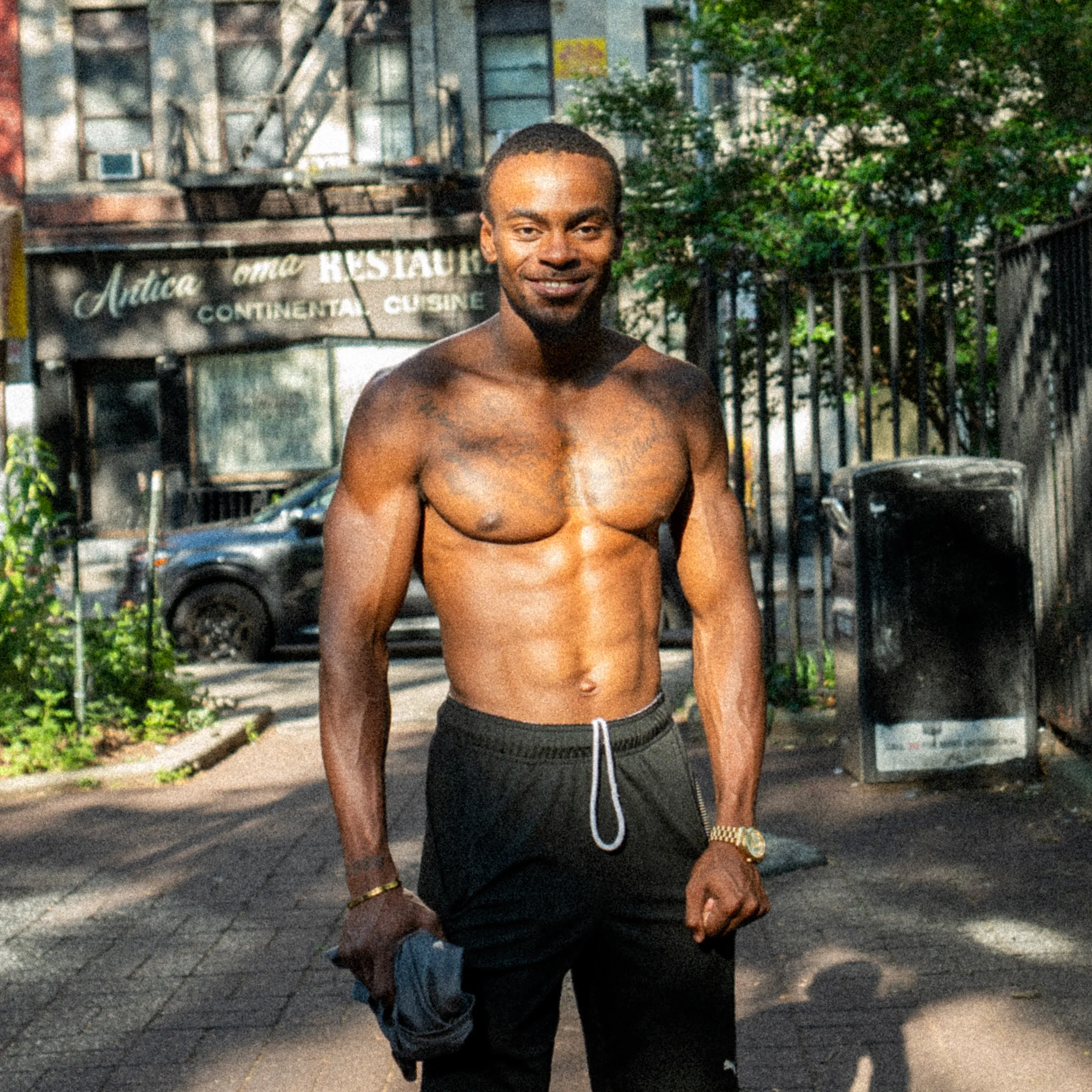 A smiling shirtless man poses on a sunlit sidewalk.