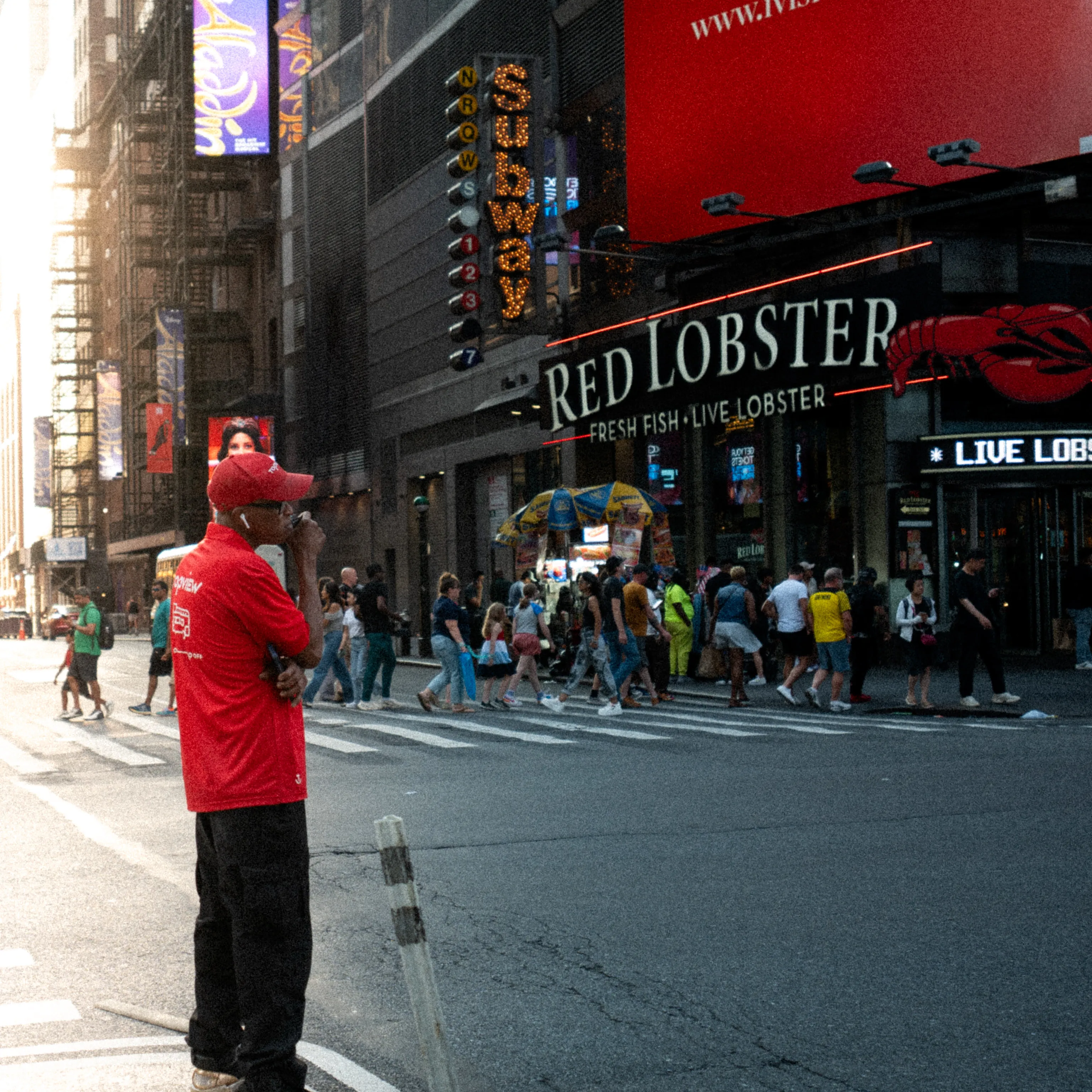 A man in a red shirt and cap stands at a Times Square crossing under the Red Lobster sign.