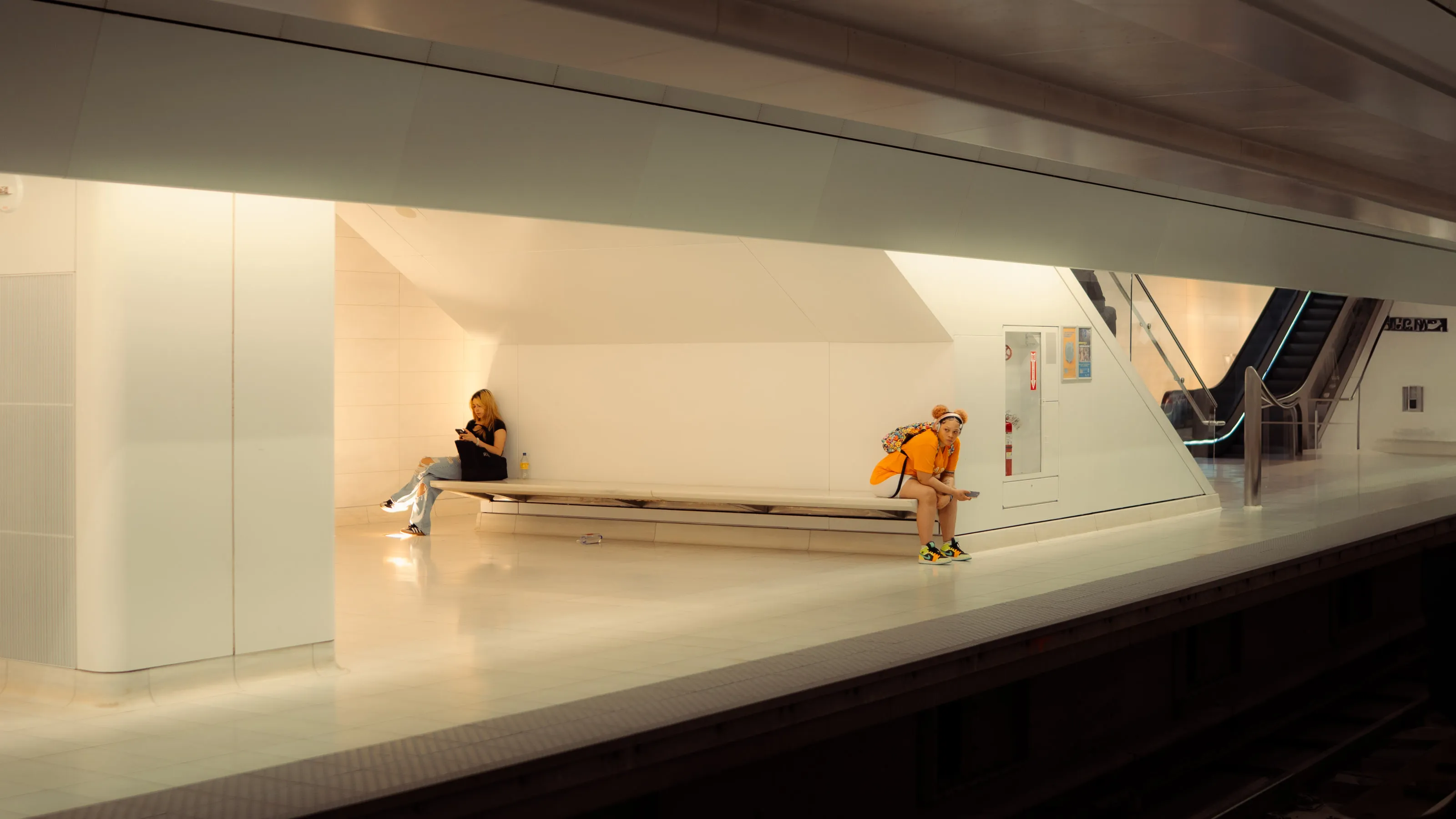 Two people wait on benches in a long, white, empty station.