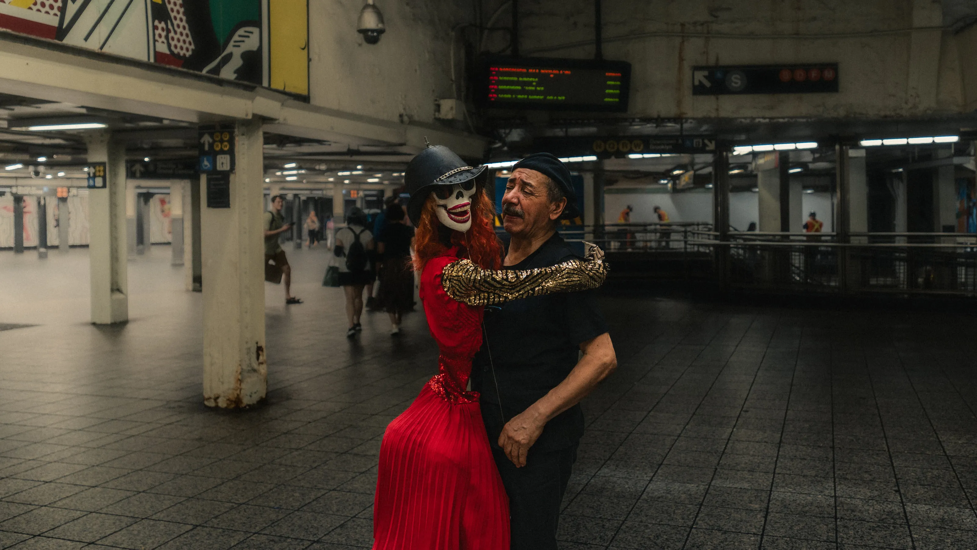 A performer in white face paint and a red dress embraces a man in a subway concourse.