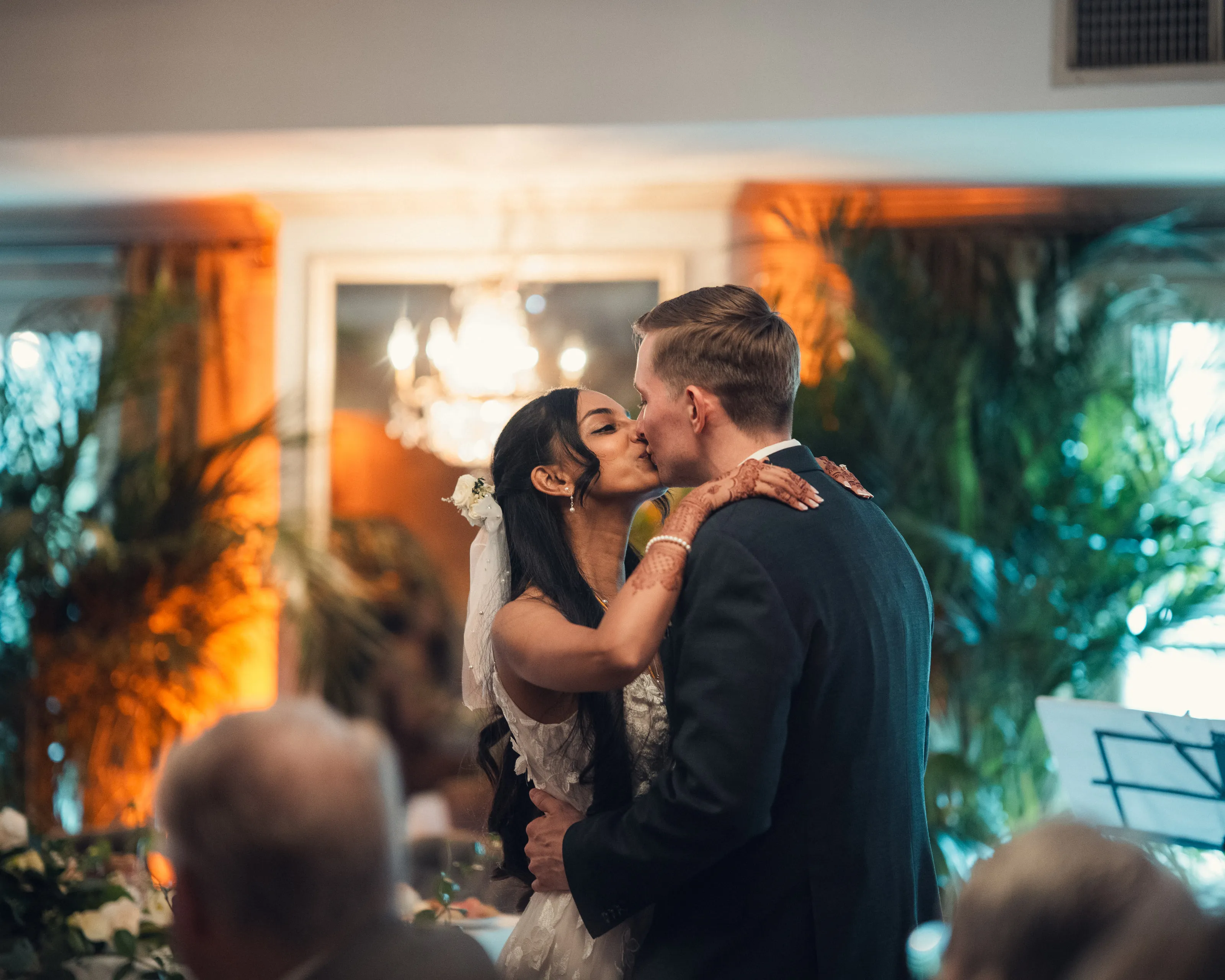 The couple kiss under warm light, with guests in the foreground.