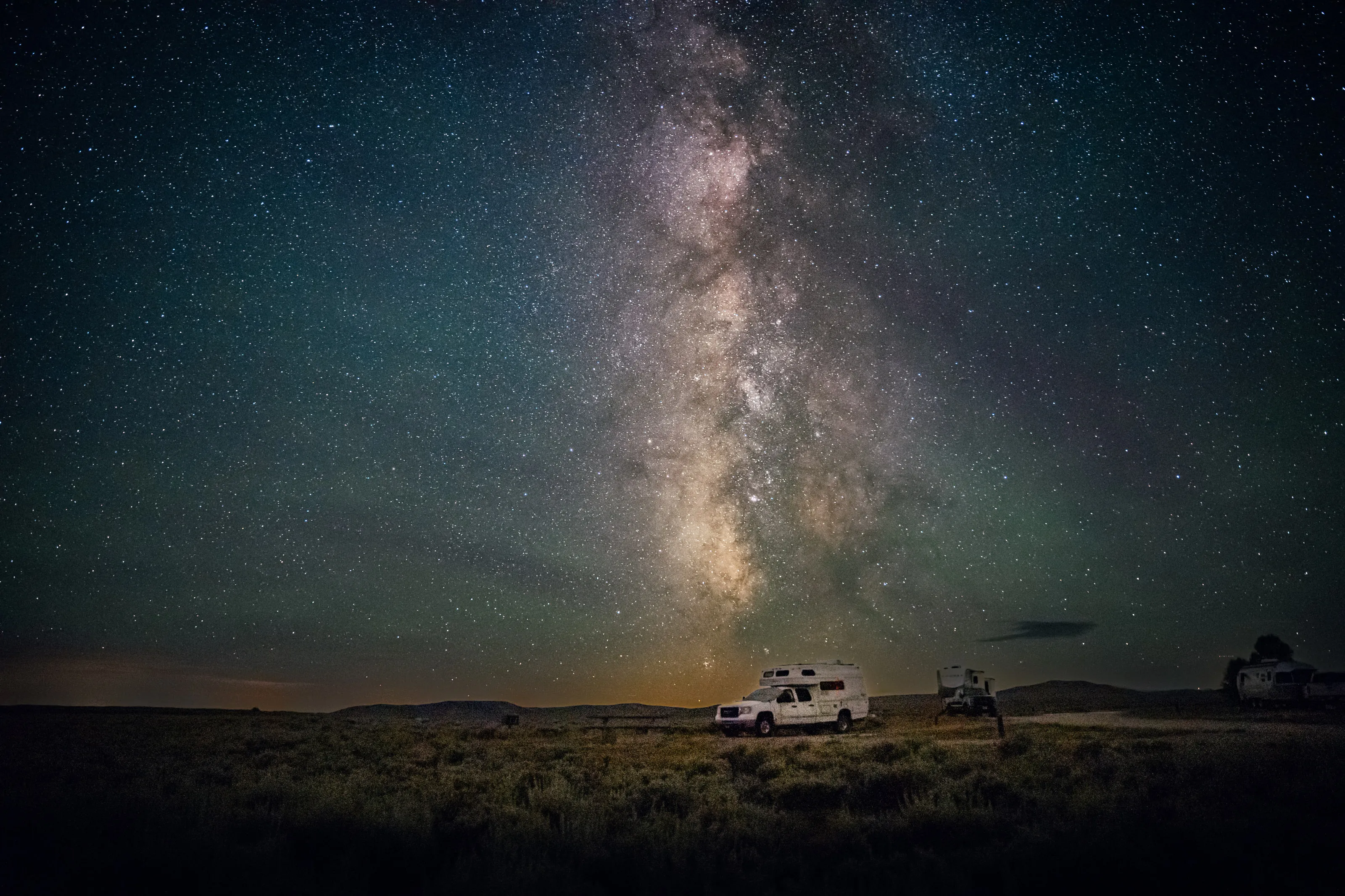 The Milky Way rises over a camper parked on the open plain at night.