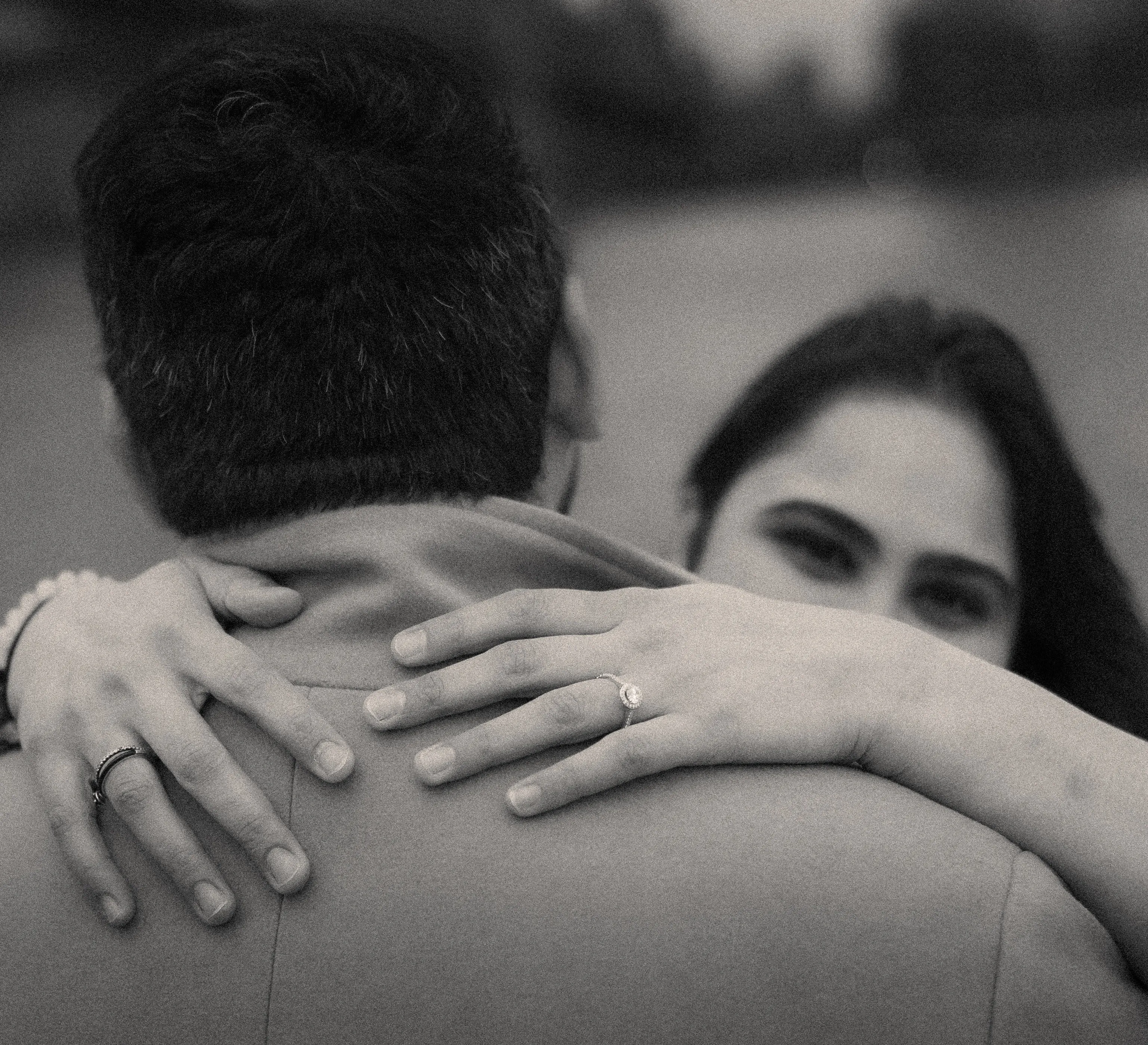 Black-and-white close-up of a woman's hand with an engagement ring on a man's shoulder.
