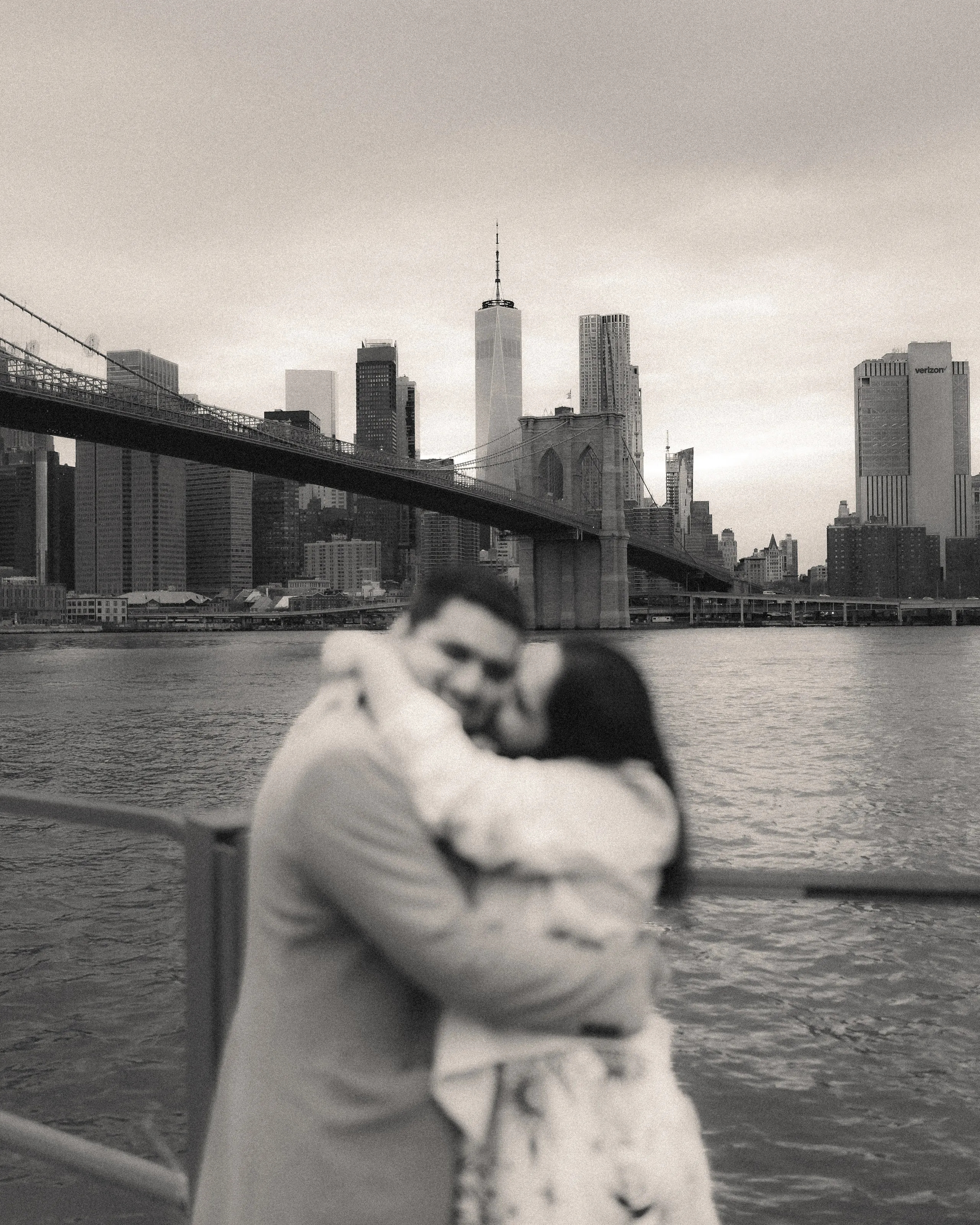 Black-and-white photograph of the couple embracing by the water, the Brooklyn Bridge behind them.