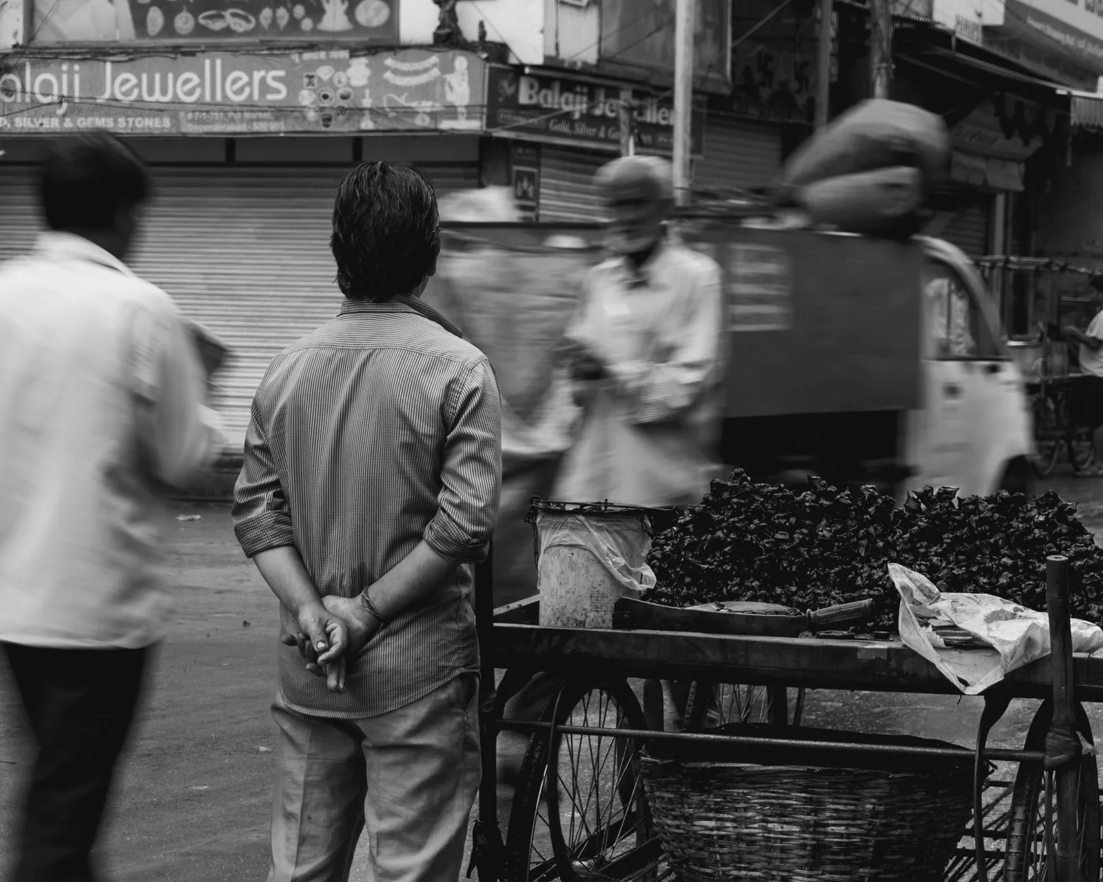 Black-and-white street scene: men watch a vendor at a cart outside a jewellery shop.
